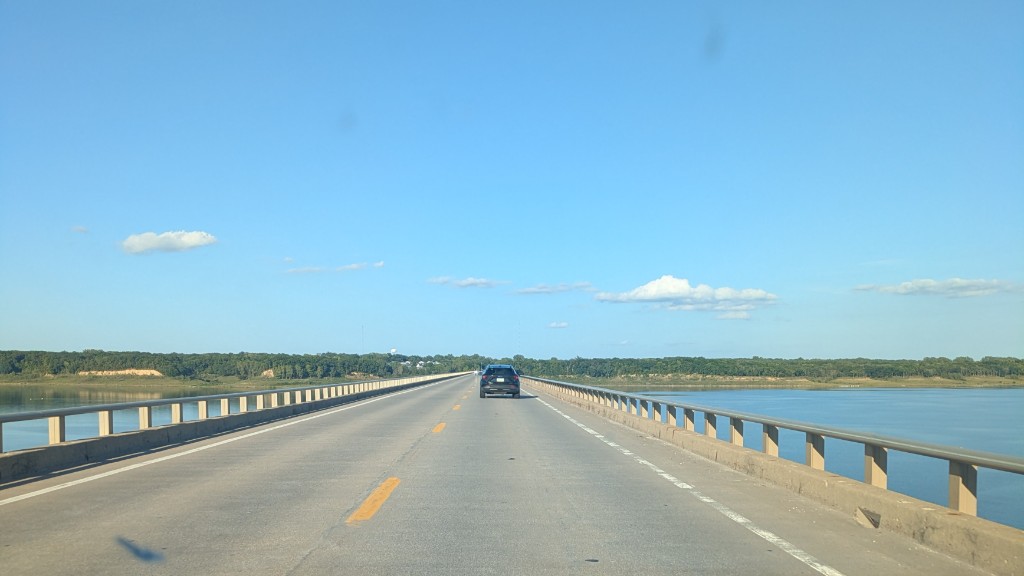 First-person photograph on a concrete causeway over water: empty lanes, yellow dashes, rails, a dark car ahead, blue sky